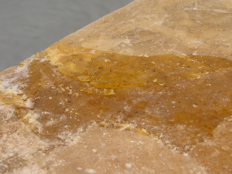 Close-up installation view of sculpture "Mediating with the rain," a large block of copal resin bound together with dust collected from various archaeological archives. The closeup centers on a small area damp with water collecting on the surface.