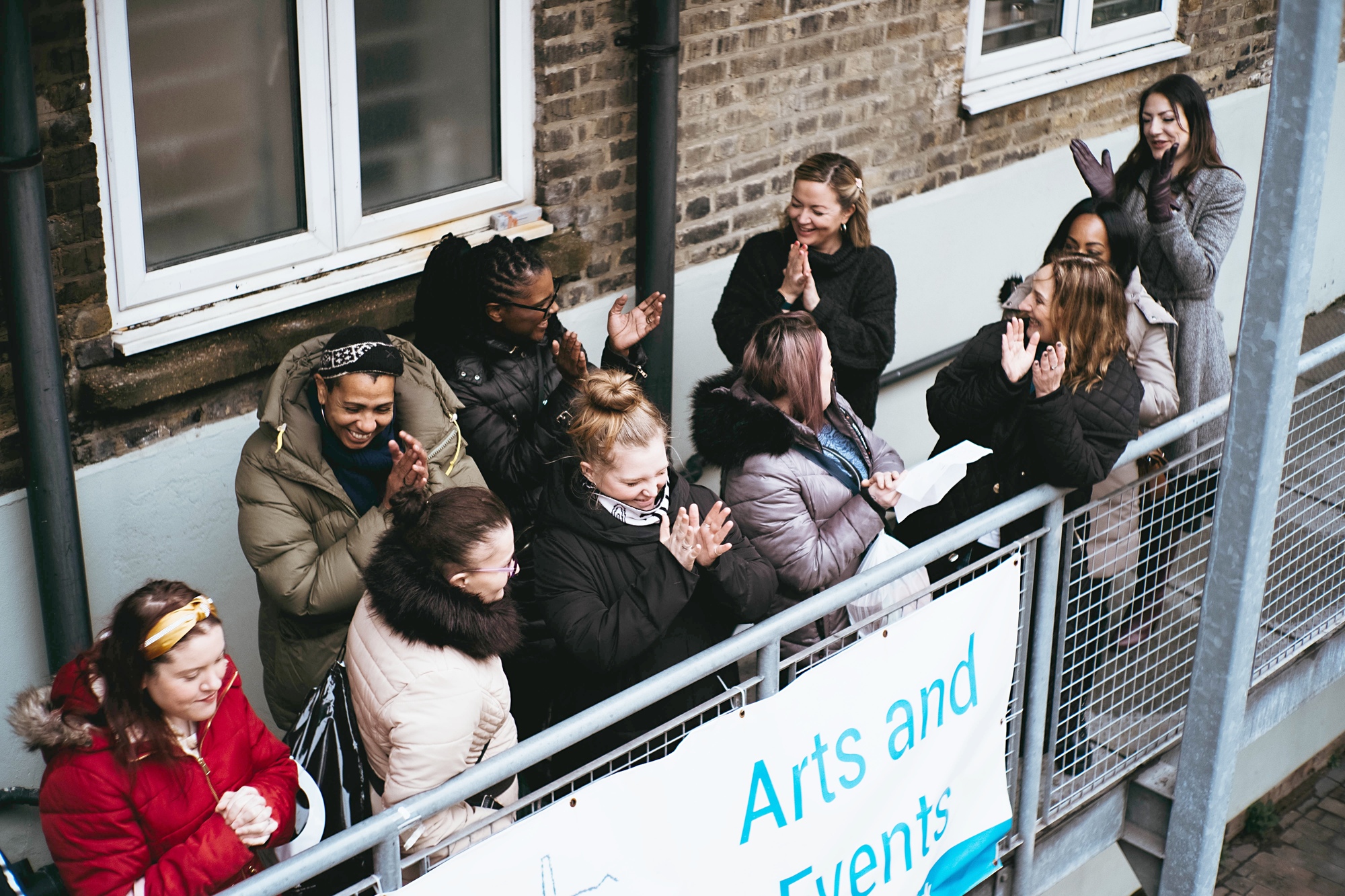 A group of women singing happily on the balcony. The women are standing close together, gazing at each other with laughter and excitement in their eyes. In front of them hangs a banner that reads Art and Event, the text is in blue.