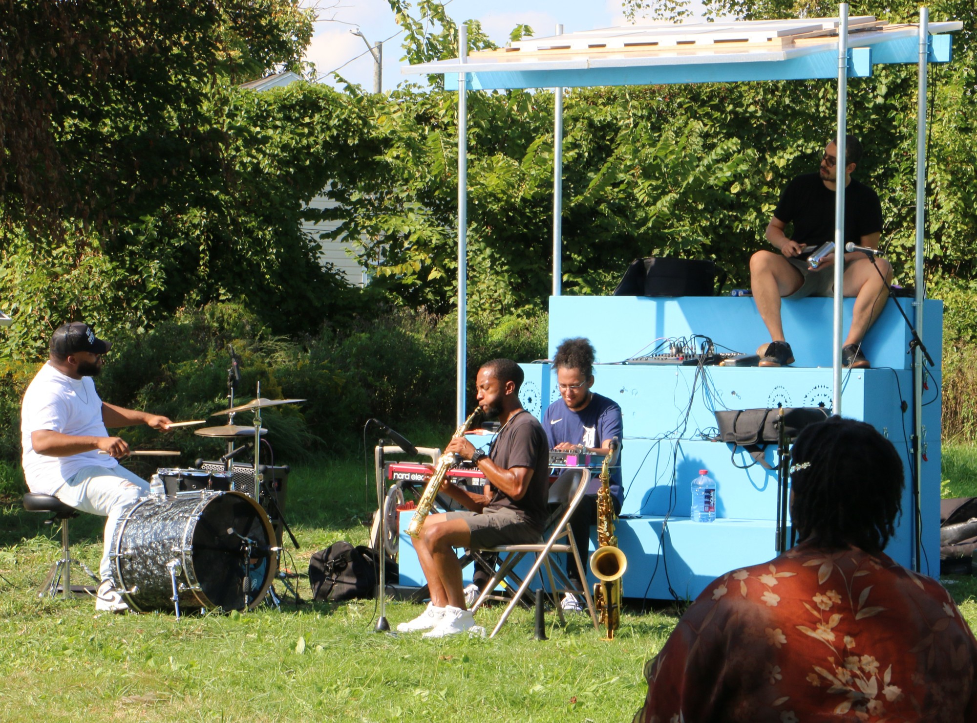 A light blue stairs-like mobile sculpture is sat on by a four-person jazz band, includes a drummer, saxophone player.